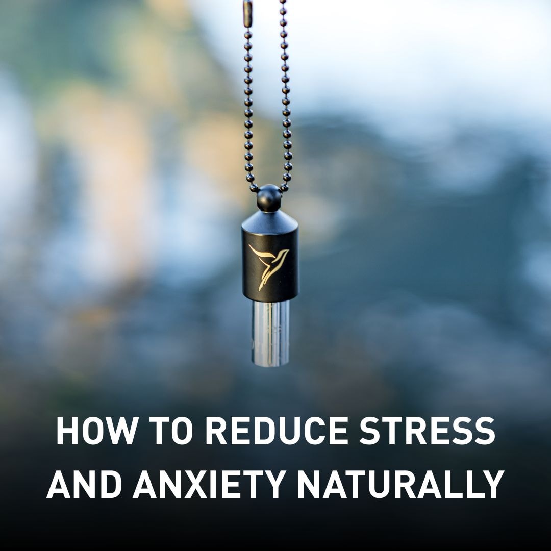 Close-up of a black breathing pendant with a hummingbird logo hanging from a beaded chain, blurred water in the background. Text overlay: 'How to Reduce Stress and Anxiety Naturally