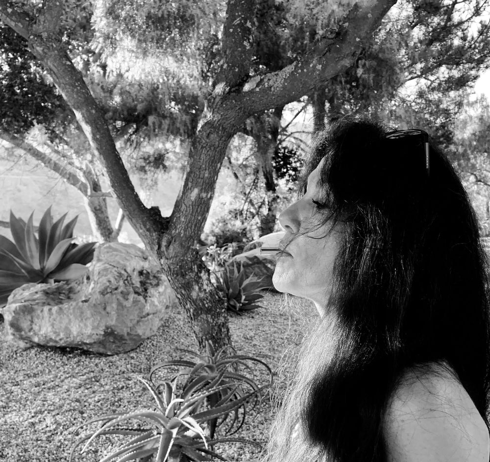 A black and white photo of a woman with dark hair using the Lovetuner. There are trees, rocks, and aloe vera plants behind her.