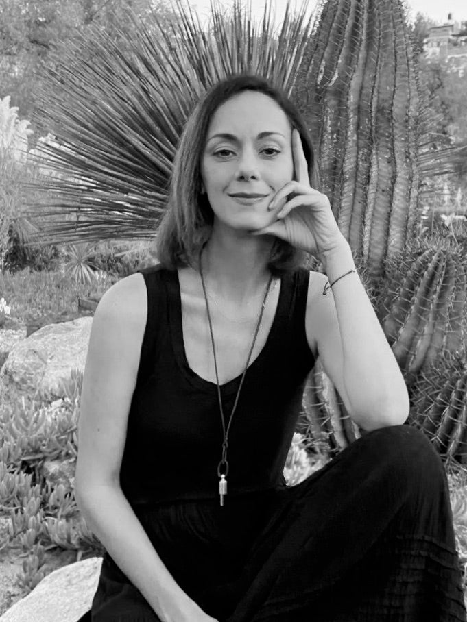 A black and white photo of a woman in a black outfit sitting amongst rocks and cacti wearing a Lovetuner necklace