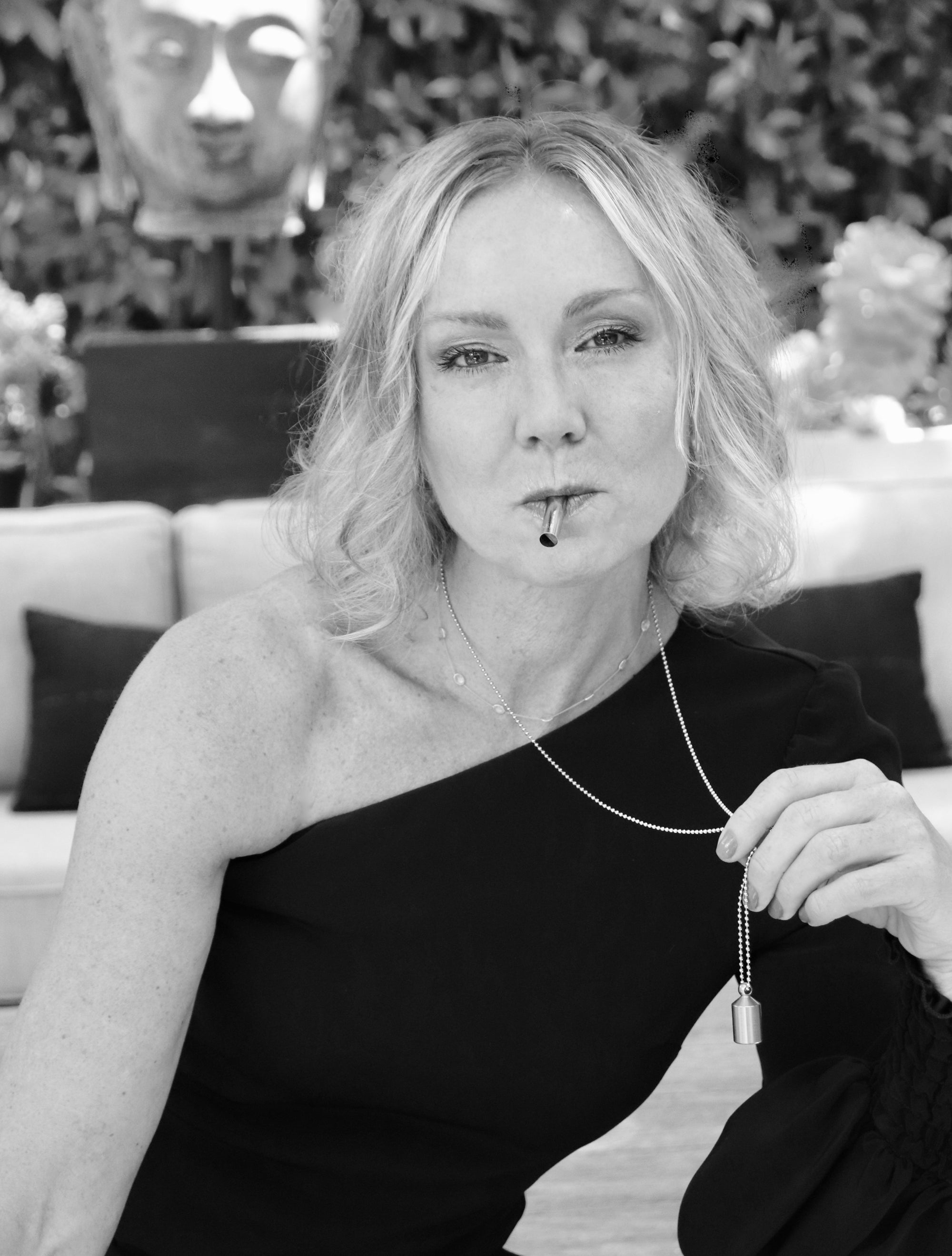 A black and white photo of a woman in a black asymmetrical off-shoulder dress using the Lovetuner and holdnig the chain with her hand with a Buddha head blurred in the background behind her.